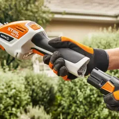 Ręka w rękawicy trzyma biało-pomarańczowe nożyce akumulatorowe Stihl HSA 26, gotowa do pracy w ogrodzie.