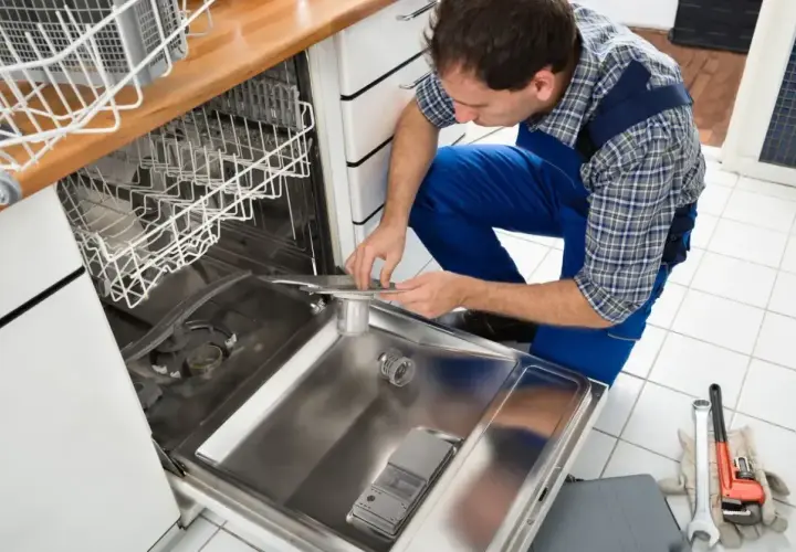 Technik w niebieskim kombinezonie pokazuje, jak podłączyć zmywarkę, pracując przy otwartych drzwiach urządzenia.
