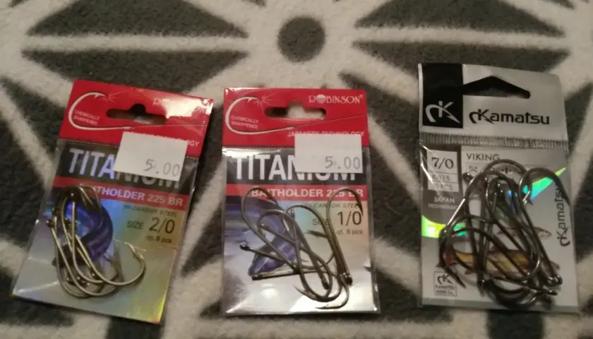 Trzy opakowania haczyków wędkarskich: dwa z napisem "TITANIUM" i jeden z logo "Kamatsu".