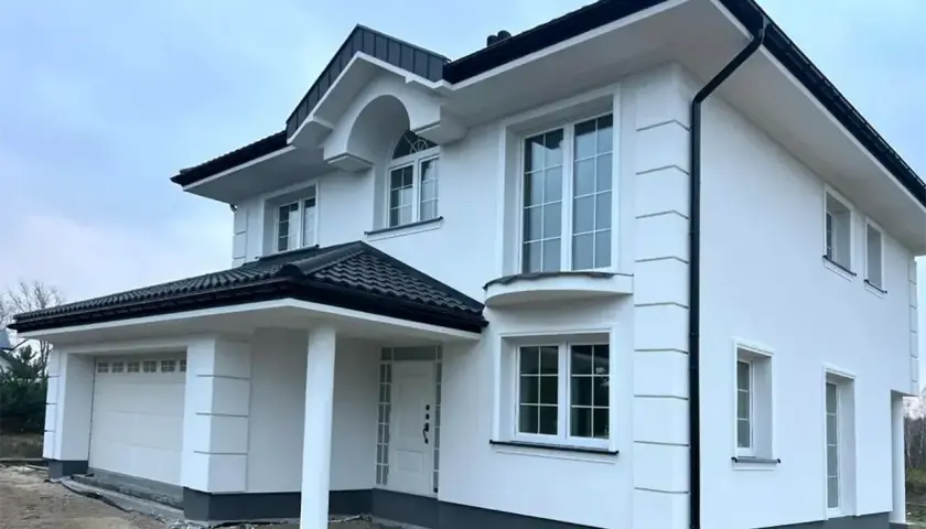 Nowy dom z białym tynkiem, gotowy do malowania elewacji. Eleganckie detale i czarny dach dodają mu uroku.