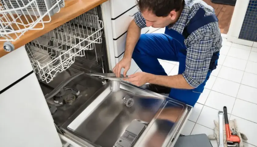 Technik w niebieskim kombinezonie pokazuje, jak podłączyć zmywarkę, pracując przy otwartych drzwiach urządzenia.