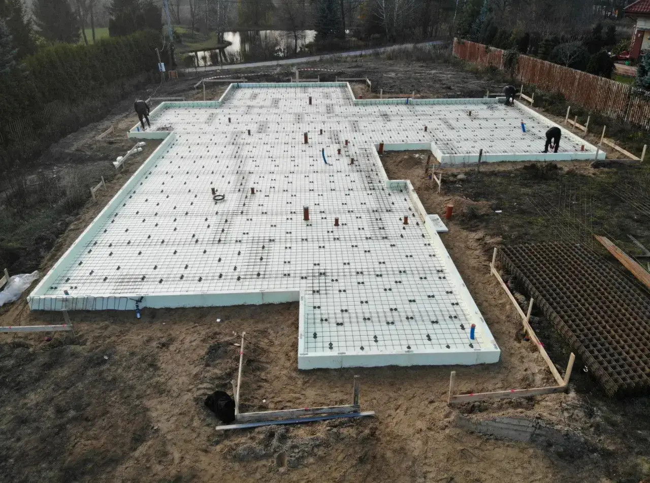 Budowa domu w toku: przygotowane stopy fundamentowe z izolacją i zbrojeniem, pracują budowlańcy.