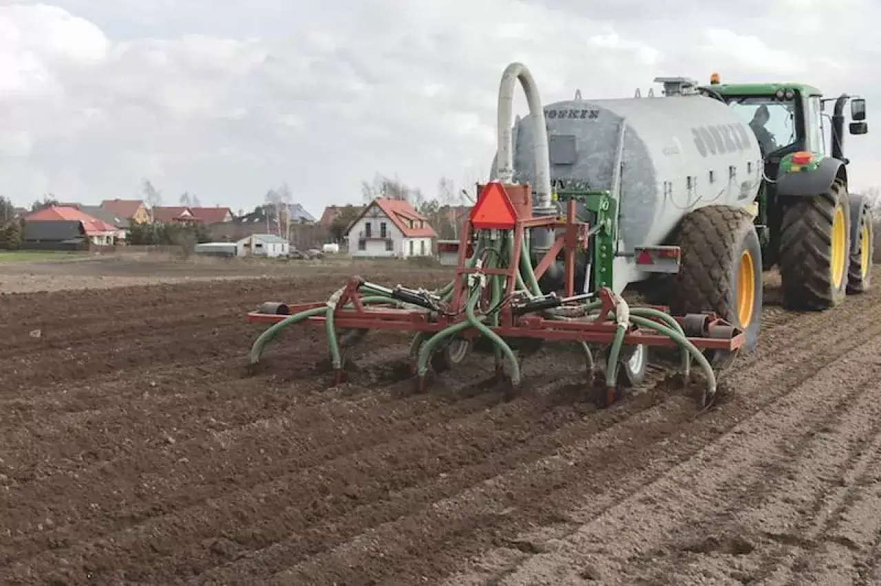 Zielony traktor z przyczepą-rozsiewaczem na polu. Maszyna rolnicza, rekultywator, przygotowuje ziemię.
